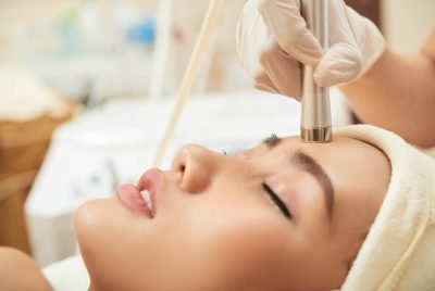 Close-up shot of relaxed young woman lying on treatment table while highly professional beautician massaging her face with special equipment